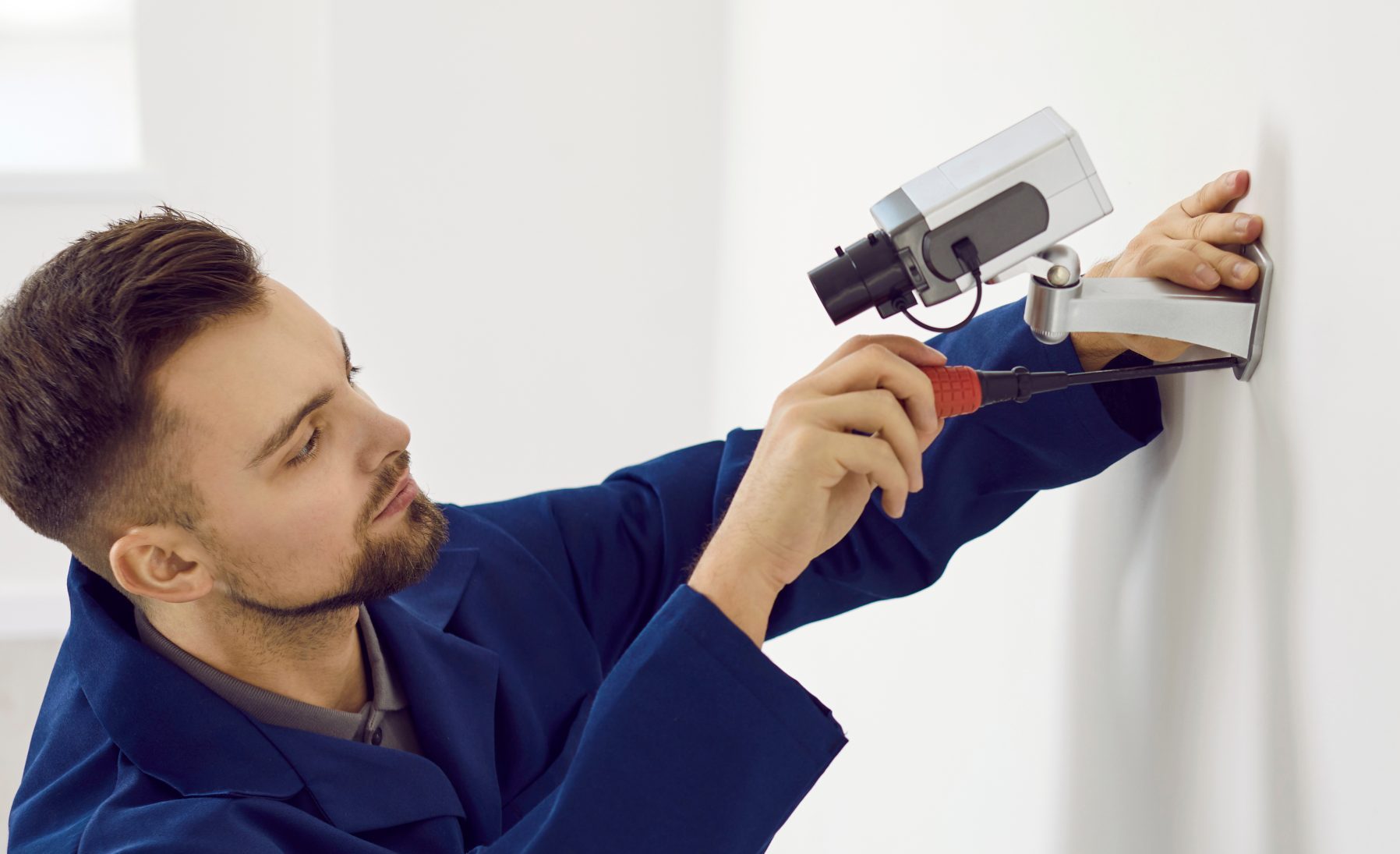 Repair service worker uses screwdriver to fit screws and adjust wall mounted security camera. Young male technician installing CCTV surveillance cam for theft deterrence at home or in office building